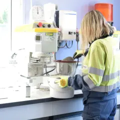 Trabajadora en laboratorio, usando guantes, manipula un recipiente con material. Se investiga qué es la silicona y sus usos en la industria.