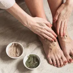 Kobieta aplikuje na stopy peeling, przygotowując je do użycia skarpetek złuszczających. Dwie miseczki z kosmetykami stoją obok.