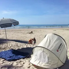 Na plaży rozłożony namiot i koc, idealne miejsce, by sprawdzić, czy można spać na plaży. Dzieci bawią się w piasku, a w tle widać morze.