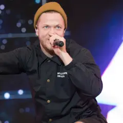 Imagine Dragons wokalista, Dan Reynolds, w beżowej czapce i czarnej kurtce, śpiewa do mikrofonu, pokazując znak pokoju.