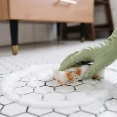 Ręka w zielonej rękawiczce czyści gąbką białe heksagonalne płytki z pianą.