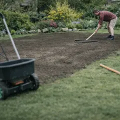 Mężczyzna zgrabia ziemię, przygotowując ją do siewu. W tle wózek rozsiewający, kluczowy element, jak wyrównać trawnik.