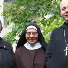 Drei Geistliche, zwei Nonnen und ein Priester, stehen lächelnd vor einem Gebäude. Sie repräsentieren katholische Orden.