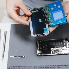 Ręce trzymające rozebrany telefon, narzędzia i niebieskie urządzenie do naprawy.