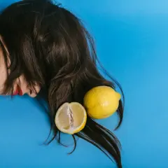 En person med mörkt hår och röda läppar ligger på en blå bakgrund. Två citroner, en hel och en delad, ligger bland håret, som om de används för att bleka håret med citron.