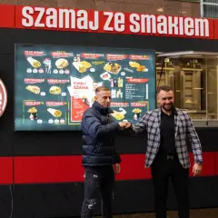 Dwóch mężczyzn wita się przy budce z kebabem, na której widnieje menu.