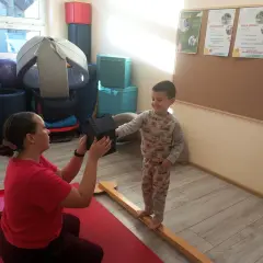 Fizjoterapeutka podaje chłopcu domek, który stoi na drewnianej belce.