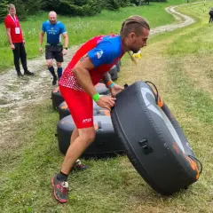 Uomo con treccia si allena al parco, spingendo un grande gonfiabile nero. Altri partecipanti corrono su un sentiero erboso.