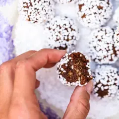 En hand håller en bit av nyttiga chokladbollar, rullade i kokosflingor, på en vacker lila-vit tallrik.