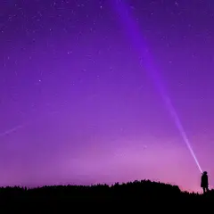 Sylwetka człowieka oświetla latarką nocne niebo, na którym widać gwiazdy i **gwiazdozbiór Tarczy**.