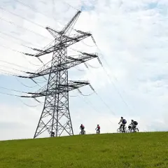 Ludzie na rowerach jadą po zielonym wzgórzu pod ogromną konstrukcją linii wysokiego napięcia.