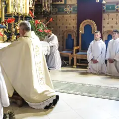 Ksiądz w szatach odprawia nabożeństwo, klęczą ministranci w białych albach.