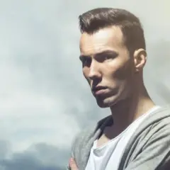 Dorian Tomasiak w szarej bluzie z kapturem i białej koszulce, z burzą chmur w tle.