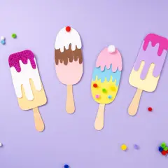 Bunte Eis-Kreationen zum Eis basteln, verziert mit Glitzer und Pompons, liegen auf lila Untergrund.