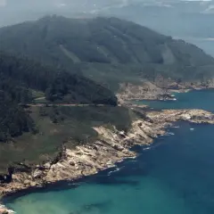 Costa rocosa y verde con el mar azul turquesa. Se ve una carretera serpenteando por la ladera. La playa Portonovo Viveiro es un paraíso natural.