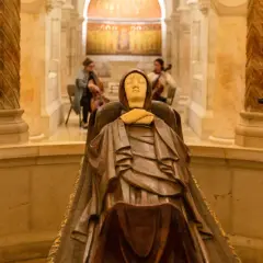Holzschnitzerei der Mutter Gottes in einer Kapelle, mit Kerzenlicht und Blumen geschmückt. Im Hintergrund spielen zwei Musikerinnen Cello.