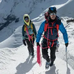 Dwóch alpinistów wspinających się po ośnieżonym zboczu, z liną i sprzętem. Zapewniają sobie asekurację lotną w trudnym terenie.