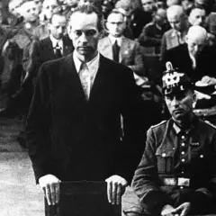 Mann im Anzug vor Gericht, umgeben von Zuschauern. Die Atmosphäre erinnert an die harten Urteile, die Roland Freisler fällte.