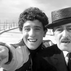 Un giovane sorridente con riccioli e un uomo anziano con cappello e baffi, un'immagine che trasmette un senso di complicità e un significato di legame generazionale.