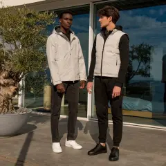 Due uomini eleganti, uno con giacca grigia e jeans, l'altro con gilet grigio e pantaloni sportivi neri, posano all'esterno.