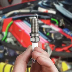 Une bougie d'allumage tenue en main, possible cause d'une voiture qui broute au démarrage. Le moteur est flou en arrière-plan.