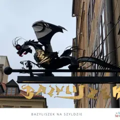 Metalowy bazyliszek ziejący ogniem wisi nad złotym napisem.