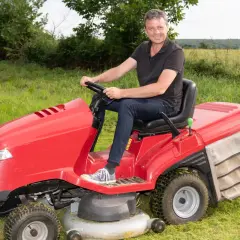 Uśmiechnięty mężczyzna siedzi za kierownicą czerwonego traktorka do koszenia trawy, gotowy do pracy w ogrodzie.