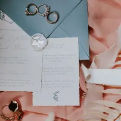 Eleganckie zaproszenia ślubne, obrączki i ozdobne pióra na różowym tle.