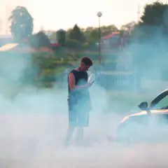 Młody mężczyzna stoi w kłębach dymu, patrząc w telefon. Obok niego samochód, z którego wydobywa się dym, jakby diesel kopcił na czarno.