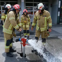 Jugendfeuerwehr übt den Umgang mit dem Hydranten. Wasser spritzt aus dem roten Standrohr, während die jungen Brandschützer in voller Montur zuschauen.