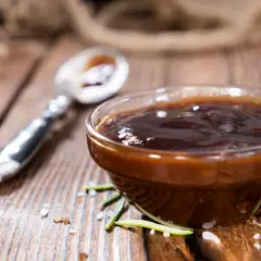 Gęsty, ciemny sos z pieczeni w szklanej miseczce, obok łyżeczka. Idealny do dań mięsnych, oto jak zrobić sos z pieczeni.