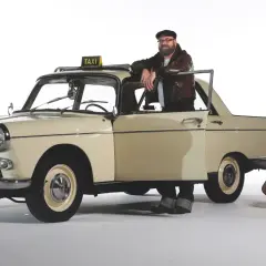 Mężczyzna w skórzanej kurtce opiera się o zabytkowy Peugeot 404 z tabliczką TAXI. Obok stoją walizki.