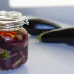 Berenjenas en escabeche, cortadas y marinadas en un frasco de vidrio, con berenjenas frescas desenfocadas al fondo.