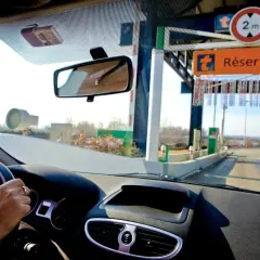 Vue depuis l'intérieur d'une voiture approchant une voie de péage. Le tableau de bord et le volant sont visibles. Un badge de télépéage est fixé au pare-brise. Le panneau "Réservé" indique comment ça fonctionne.