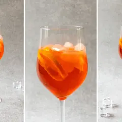 Trzy identyczne kieliszki z pomarańczowym drinkiem Aperol Spritz, lodem i plasterkami pomarańczy na szarym tle.