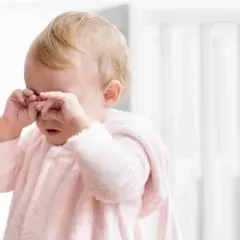 Un neonato con un pigiamino rosa strofina gli occhi, forse un occhio che lacrima per la stanchezza o un piccolo pianto.