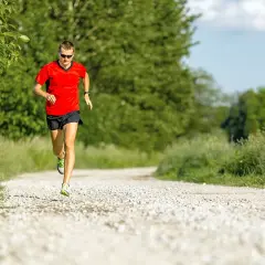 Mann in rotem Shirt und schwarzen Shorts absolviert einen Fartlek Lauf auf einem Schotterweg, umgeben von grüner Natur.