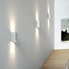 Nowoczesne oświetlenie nocne korytarza tworzy nastrojową atmosferę. Trzy białe kinkiety rzucają snopy światła na szarą ścianę, oświetlając schody i abstrakcyjny obraz.