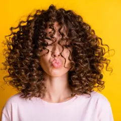Mujer con un espectacular pelo rizado fino y castaño, haciendo un beso contra un fondo amarillo vibrante.