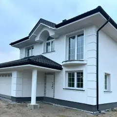 Nowy dom z białym tynkiem, gotowy do malowania elewacji. Eleganckie detale i czarny dach dodają mu uroku.