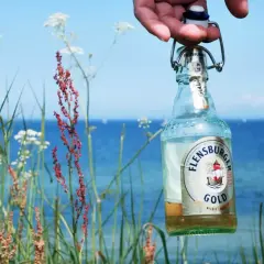 Eine Flensburger Gold Flasche wird vor der Küste gehalten. Der leichte Bier Geschmack passt perfekt zum Sommer.