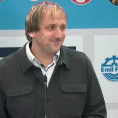 Thomas Neubert lächelt in die Kamera, trägt eine dunkle Jacke und ein gemustertes Hemd. Im Hintergrund sind Logos zu sehen.