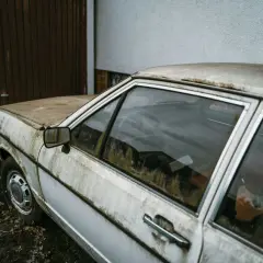 Vieille voiture blanche, couverte de poussière et de mousse, devant un garage. Un symbole de l'interdiction voiture thermique.