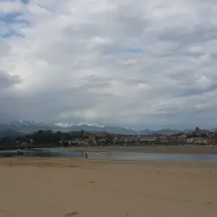 Arena dorada en la playa de la Maza, con un velero rojo en el agua y un pueblo costero al fondo, bajo un cielo nublado.