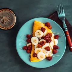 Słodki omlet z bananami i malinami, polany syropem, obok filiżanka kawy. Idealny na słodki początek dnia.