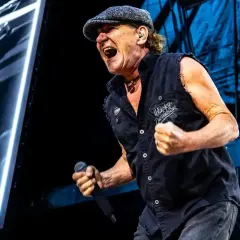 Brian Johnson z zespołu AC/DC na koncercie, w czapce i kamizelce Harley-Davidson, śpiewa z pasją do mikrofonu.