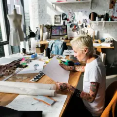 A designer sketches at a table filled with fabric and patterns, a scene that captures the essence of what is an artist residency: a dedicated space for creative exploration and development.