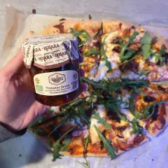 Mano sostiene un tarro de tomates secos en aceite de oliva virgen extra, listo para añadir a una pizza con rúcula.