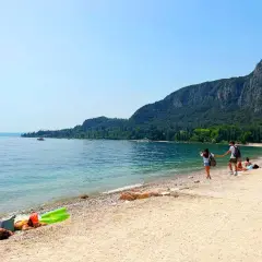 Ein Paar hält Händchen am Strand, während andere am Gardasee schwimmen. Berge und Bäume säumen die Küste.