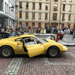 Żółte Ferrari Dino z otwartymi drzwiami, kierowca w środku, tłum widzów za barierką.